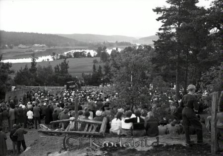 Allsångskväll på bygdegården sommaren 1955.
raja
 En samling människor framför en sångare och en man som spelar piano på platsen där idag kaffestugan ligger på Hembygdsgården i Kisa. I bakgrunden Värgårdsudde och Kisasjön. 

Nyckelord: Allsångskväll bygdegården